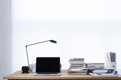 A light-colored desk with an open laptop, a desk lamp and stacks of books.