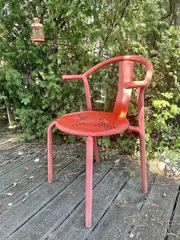 A red metal chair with slender legs and curved arms on an outdoor wooden deck.